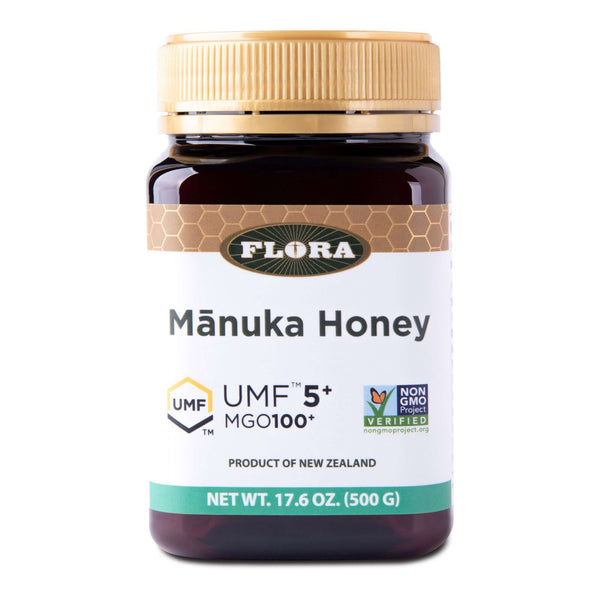 A jar of Flora Mānuka Honey with labels indicating UMF 5+ and MGO 100+ certifications, as well as being non-GMO and sustainable. The jar is brown with a gold lid and has information stating it is a product of New Zealand.