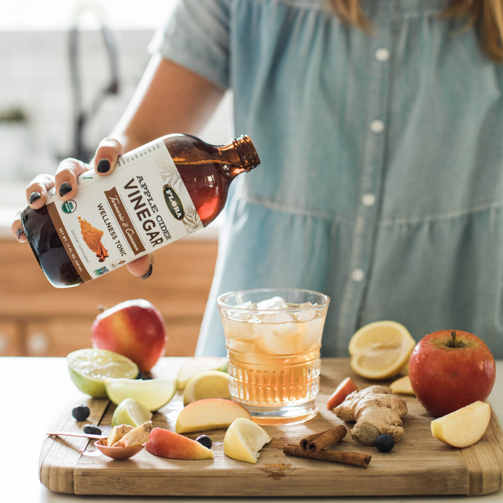 organic, non-gmo apple cider vinegar with turmeric and cinnamon poured into glass of ice surrounded by fresh fruit and herbs: apples, ginger, lemons, limes, elderberries, and ginger