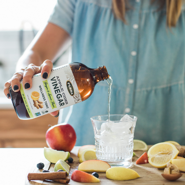 lemon ginger apple cider vinegar being poured into glass of water surrounded by fresh fruit and herbs: cinnamon, apples, lemons, limes, and elderberries